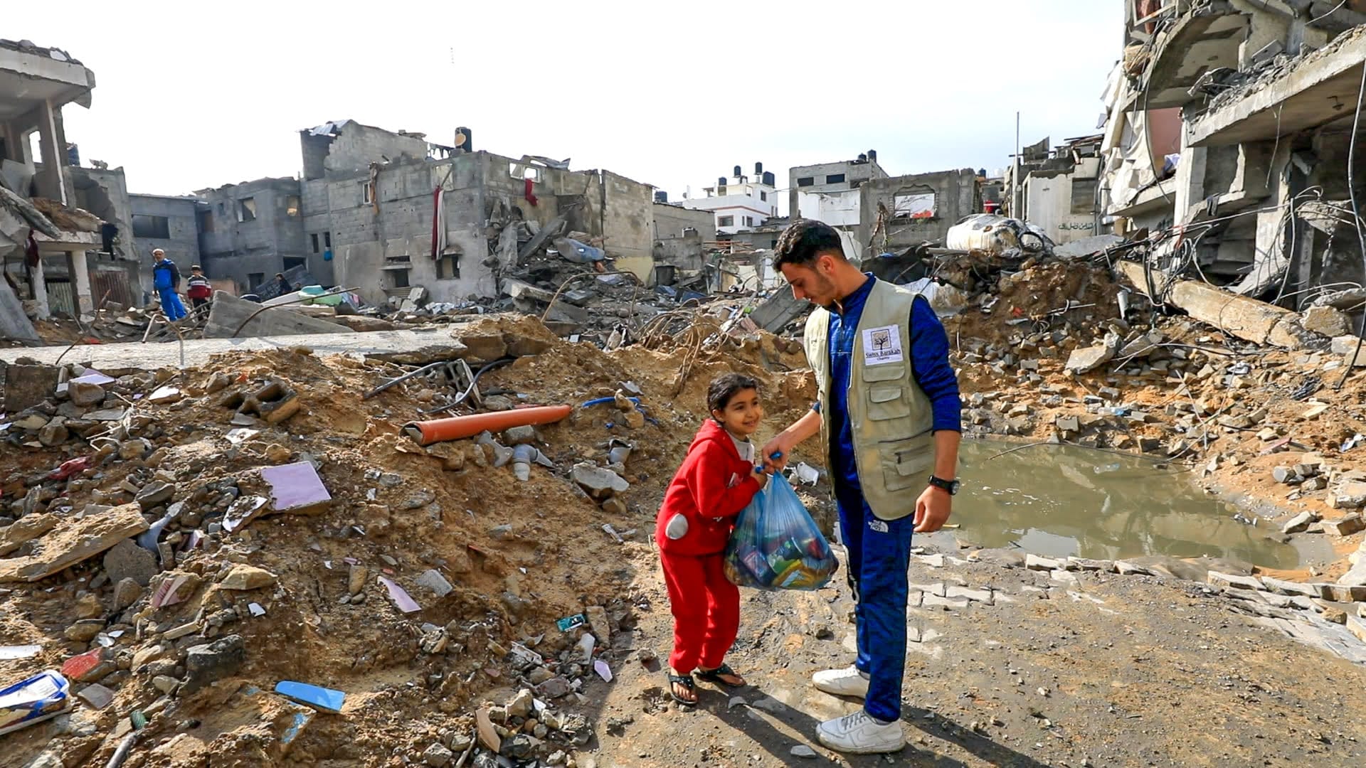 A humanitarian worker hands a bag of food supplies to a smiling girl in a red tracksuit amidst destroyed buildings.